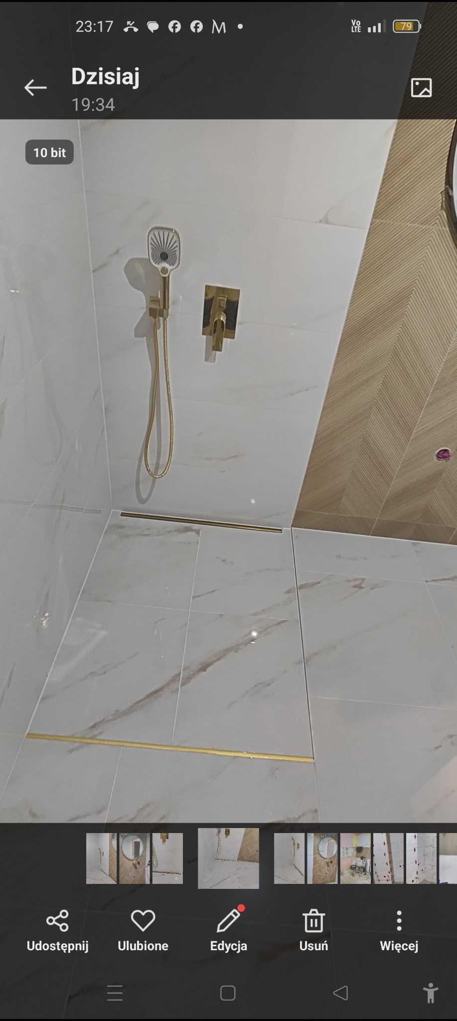 Kabina prysznicowa z białymi płytkami imitującymi marmur, złotą armaturą i odpływem liniowym, oraz fragmentem ściany wykończonej drewnopodobną płytką ułożoną w jodełkę.