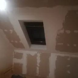 Poddasze w trakcie remontu, widoczne fragmenty ścian i sufitu z nałożoną gładzią szpachlową, okno dachowe z ciemną ramą, ślady wilgoci przy podłodze.