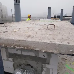 Pracownik w żółtym kasku na płaskim dachu z betonową konstrukcją, widoczne kominy wentylacyjne i elementy instalacji, w tle budynki w lekkiej mgle.