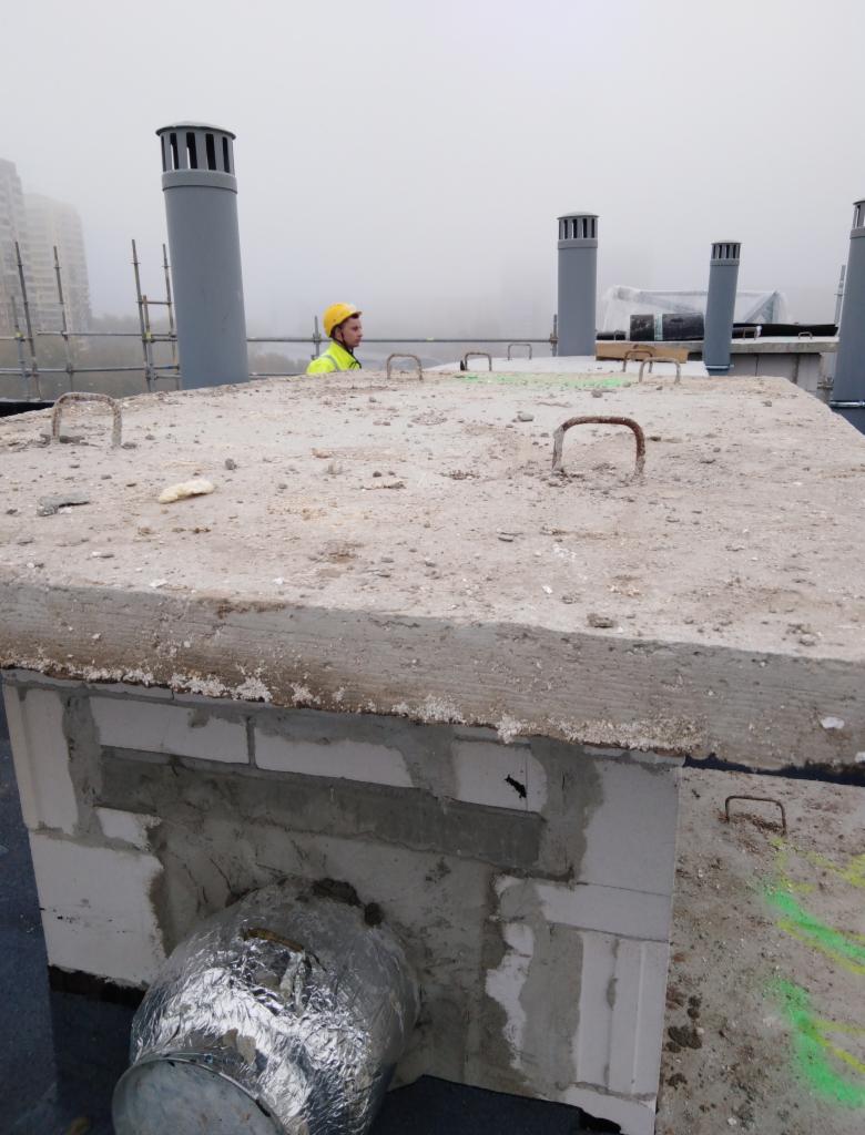 Pracownik w żółtym kasku na płaskim dachu z betonową konstrukcją, widoczne kominy wentylacyjne i elementy instalacji, w tle budynki w lekkiej mgle.