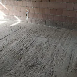 Surowy stan wnętrza: ceglana ściana z zamontowanymi puszkami elektrycznymi i poprowadzonym kablem, betonowa posadzka z widocznymi śladami szlifowania.