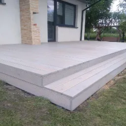 Jasnoszary taras z desek kompozytowych, dwustopniowe schody, fragment ściany budynku z elementami dekoracyjnymi z kamienia, huśtawka ogrodowa w tle.