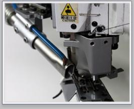 Fragment maszyny szwalniczej z metalowym ramieniem i ostrzeżeniem o zagrożeniu, skierowany w dół mechanizm dociskowy na metalowej platformie.