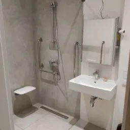 Wyremontowana łazienka z udogodnieniami dla osób niepełnosprawnych: uchwyty przy umywalce i pod prysznicem, składane siedzisko pod prysznicem, odpływ liniowy w podłodze, biała umywalka i szara...