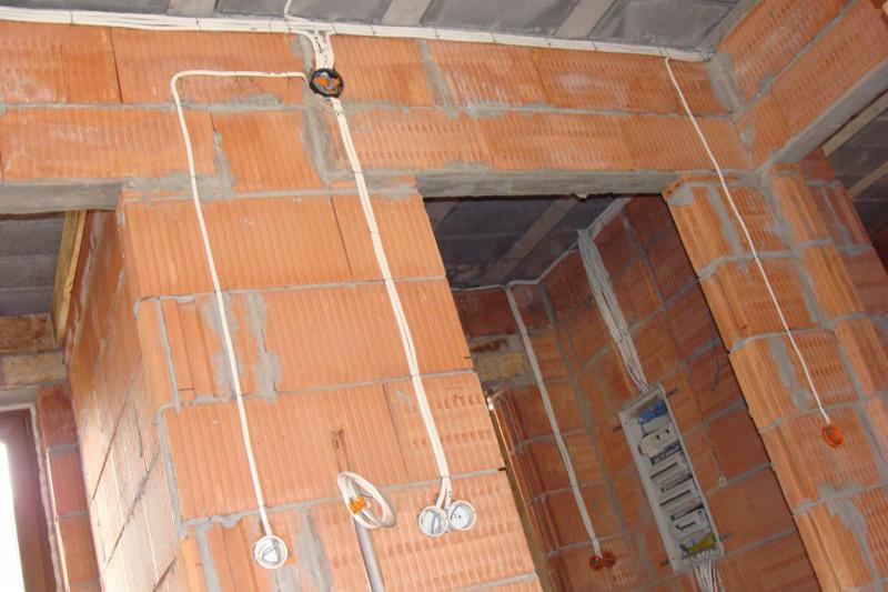 Surowy mur z cegły dziurawki z poprowadzonymi przewodami elektrycznymi, widoczna skrzynka rozdzielcza i puszki pod gniazdka, instalacja w trakcie realizacji.