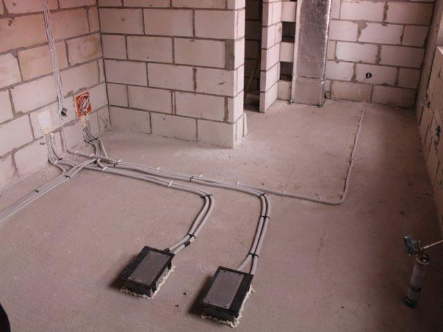 Surowy stan pomieszczenia z widocznymi przewodami elektrycznymi prowadzonymi po podłodze i ścianach, puszki instalacyjne w ścianie, szare kable przymocowane do podłoża, pianka montażowa wokół...