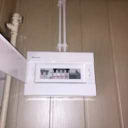 Biała skrzynka elektryczna z bezpiecznikami marki Elektro-Plast zamontowana na ścianie z panelami, z otwartymi drzwiczkami, widoczne przewody w białej rurce karbowanej.