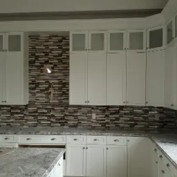 Biała kuchnia z mozaikową ścianą nad blatem, górne szafki z przeszklonymi frontami, widoczne niedokończone podłączenie oświetlenia.