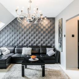 Cristal-Bud - Nowoczesny salon na poddaszu z czarną skórzaną sofą narożną, geometryczną ścianą 3D w odcieniach szarości, srebrnym żyrandolem i czarnym stolikiem kawowym z owocami.