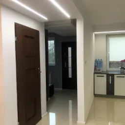 Nowoczesny przedpokój z brązowymi drzwiami, białymi ścianami i podświetleniem LED w suficie oraz błyszczącą podłogą z płytek.