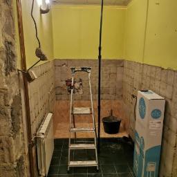 Cristal-Bud - Łazienka w trakcie demontażu płytek, widoczny grzejnik, drabina, wiadro, laser krzyżowy na statywie i karton z nową armaturą.