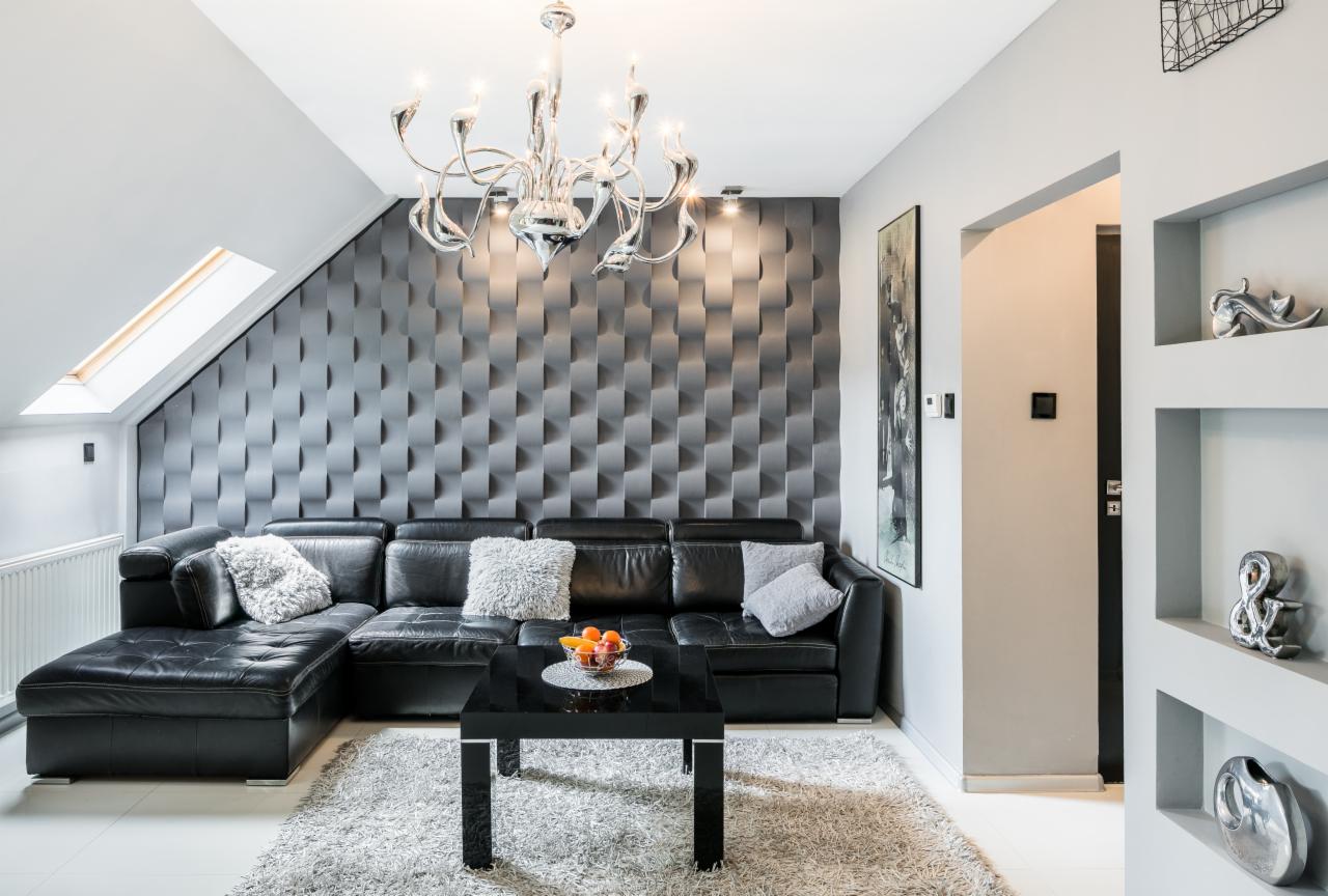 Nowoczesny salon na poddaszu z czarną skórzaną sofą narożną, geometryczną ścianą 3D w odcieniach szarości, srebrnym żyrandolem i czarnym stolikiem kawowym z owocami.