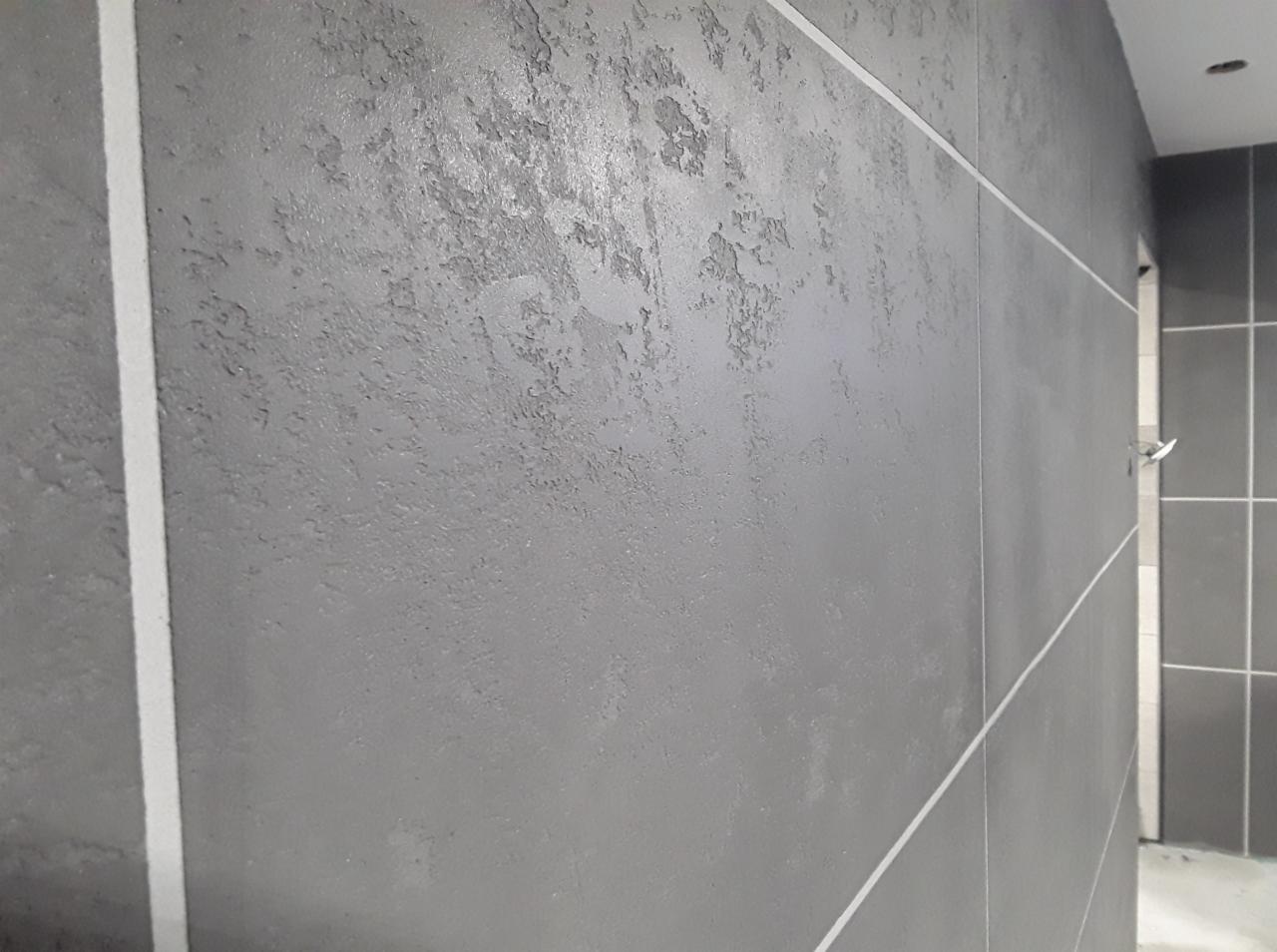 Szara ściana z efektem dekoracyjnym przypominającym beton architektoniczny, oddzielona białymi listwami, widok na fragment łazienki z płytkami w tle.