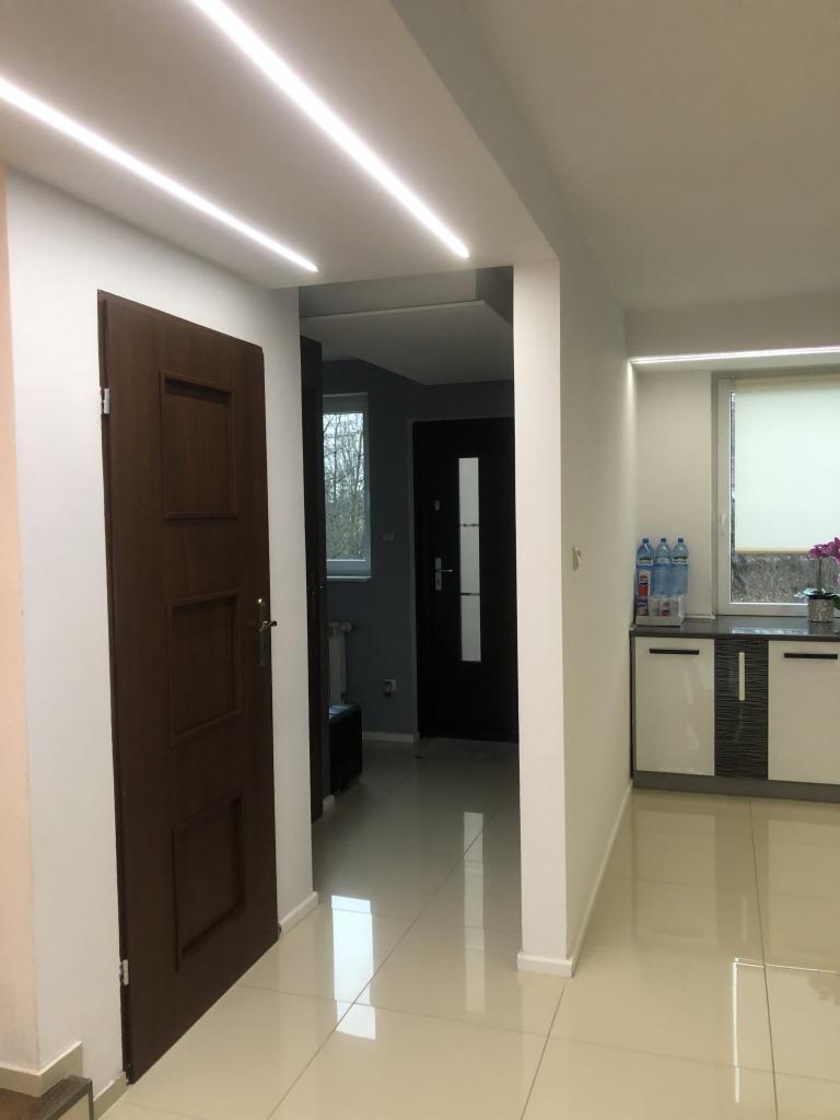 Nowoczesny przedpokój z brązowymi drzwiami, białymi ścianami i podświetleniem LED w suficie oraz błyszczącą podłogą z płytek.