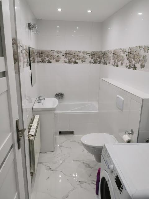 Jasna łazienka z marmurową podłogą, wzorzystymi płytkami na ścianach, białą wanną, umywalką, toaletą i pralką, widziana z perspektywy drzwi.