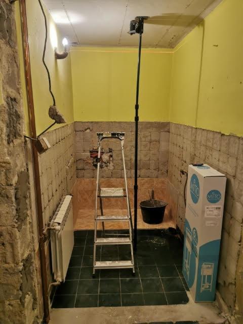 Łazienka w trakcie demontażu płytek, widoczny grzejnik, drabina, wiadro, laser krzyżowy na statywie i karton z nową armaturą.