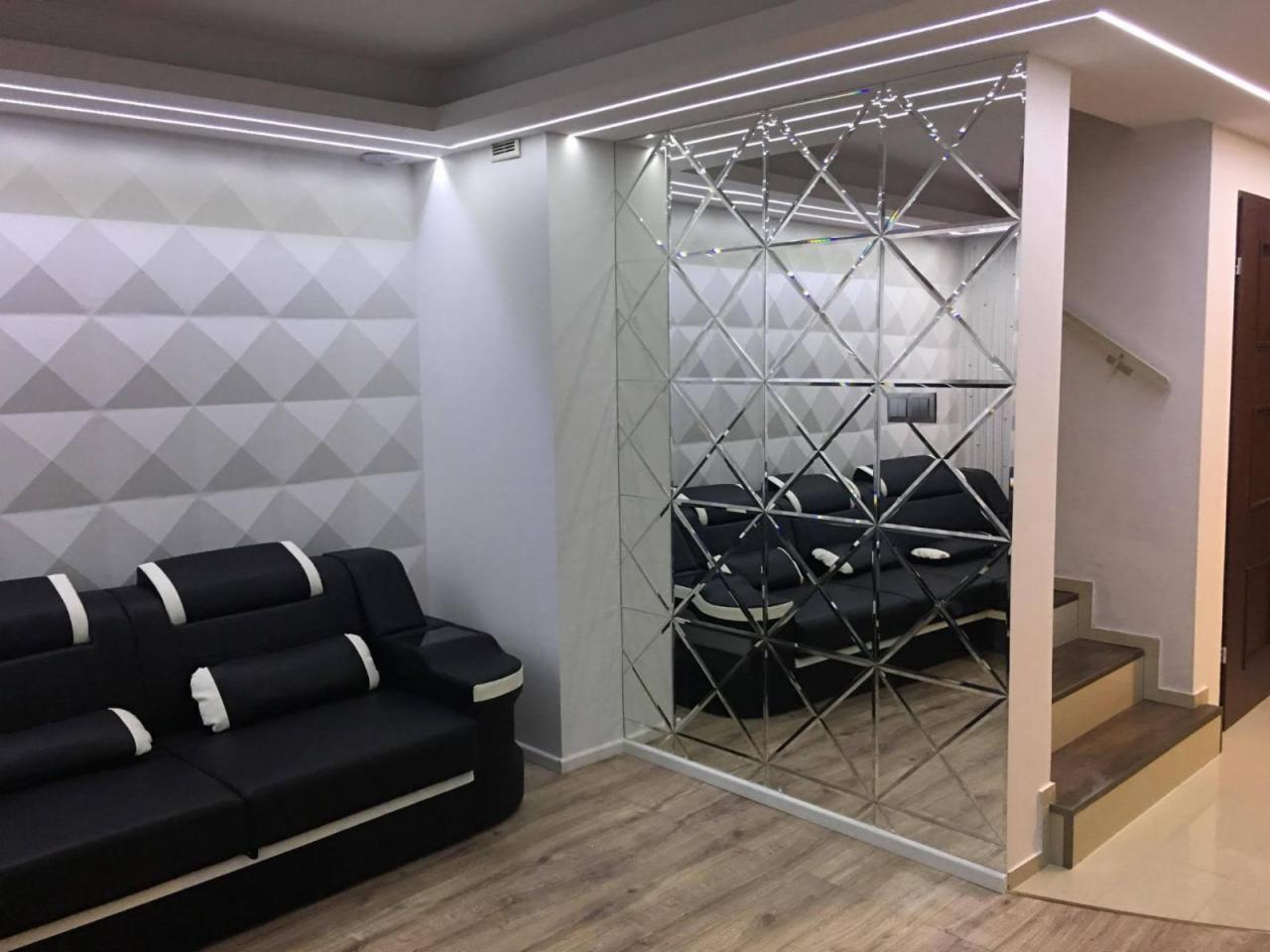 Salon z lustrzaną ścianą z geometrycznym wzorem, oświetleniem LED podsufitowym i czarną sofą z białymi akcentami, widoczne schody w tle.