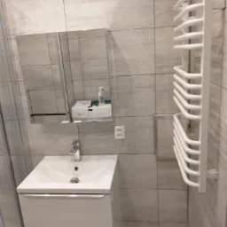 Nowoczesna łazienka z białym zestawem umywalkowym z szafką, grzejnikiem drabinkowym na ścianie z szarych płytek imitujących kamień oraz lustrem, w którym odbija się pralka i środki czystości.