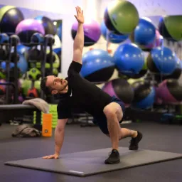 Mężczyzna w czarnej koszulce i granatowych spodenkach wykonuje ćwiczenie rozciągające na szarej macie w siłowni, z piłkami do ćwiczeń w tle.