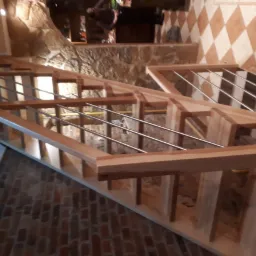 Surowe drewniane schody z metalowymi prętami balustrady, w trakcie montażu, na tle kamiennej ściany i podłogi z cegły.