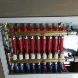 Rozdzielacz ogrzewania podłogowego w skrzynce, z widocznymi rurami w kolorach czerwonym i niebieskim, oraz zaworami i przepływomierzami.