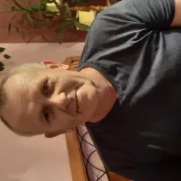 Portret mężczyzny z bliska, z delikatnym uśmiechem, na tle ściany w odcieniach różu i zieleni. Widoczne elementy dekoracyjne, ciepłe oświetlenie.