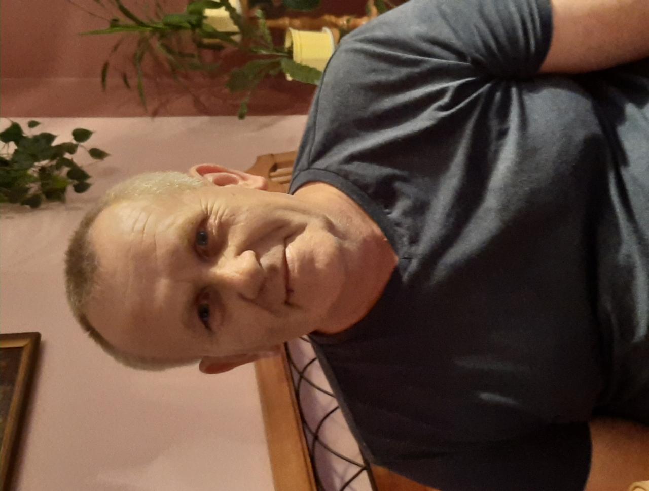 Portret mężczyzny z bliska, z delikatnym uśmiechem, na tle ściany w odcieniach różu i zieleni. Widoczne elementy dekoracyjne, ciepłe oświetlenie.