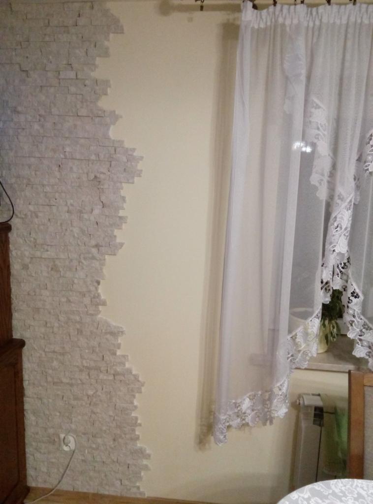 Fragment ściany z dekoracyjną okładziną z jasnego kamienia, przylegający do gładkiej, pomalowanej ściany; obok okno zasłonięte firanką z koronkowym wykończeniem.