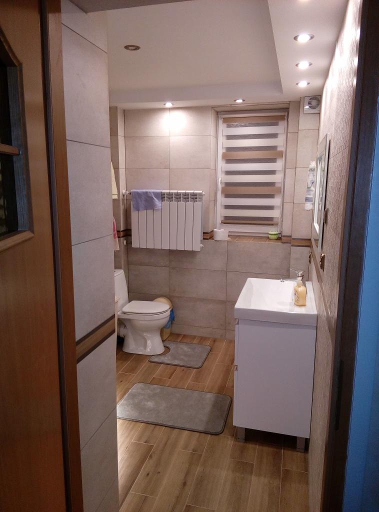 Ukończona łazienka z beżowymi płytkami, drewnopodobną podłogą, białą umywalką z szafką, toaletą i grzejnikiem pod oknem z roletami.