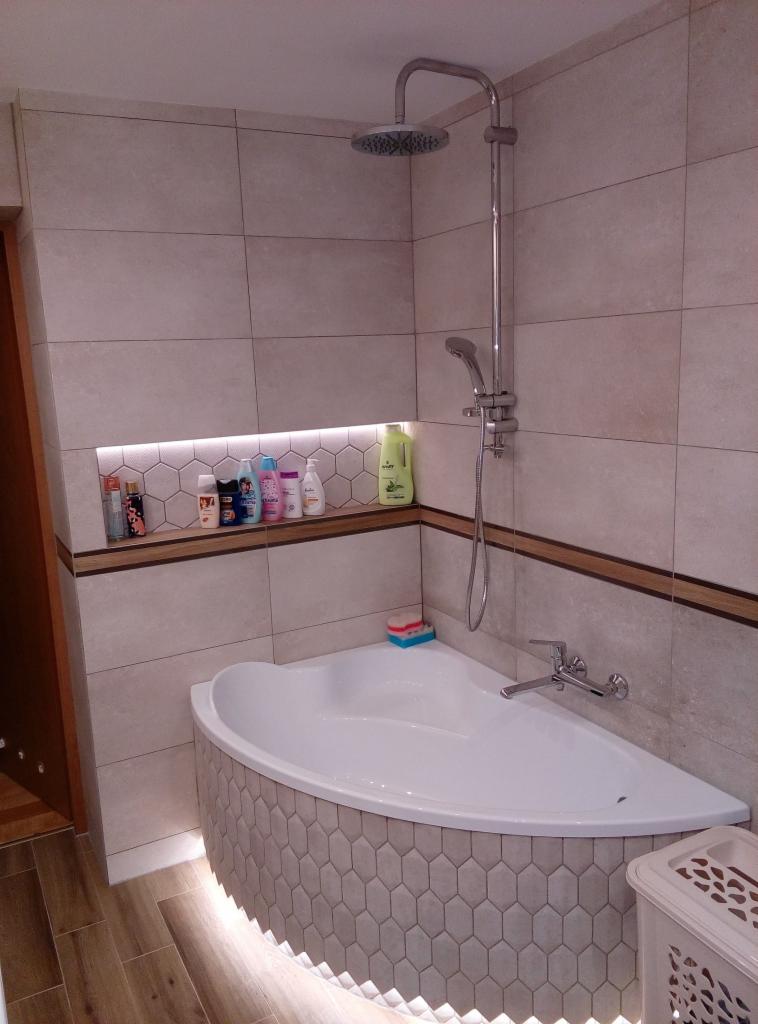 Narożna wanna obłożona heksagonalnymi płytkami z podświetleniem LED, półka ścienna z oświetleniem i kosmetykami, beżowe płytki ścienne, drewnopodobna listwa dekoracyjna.