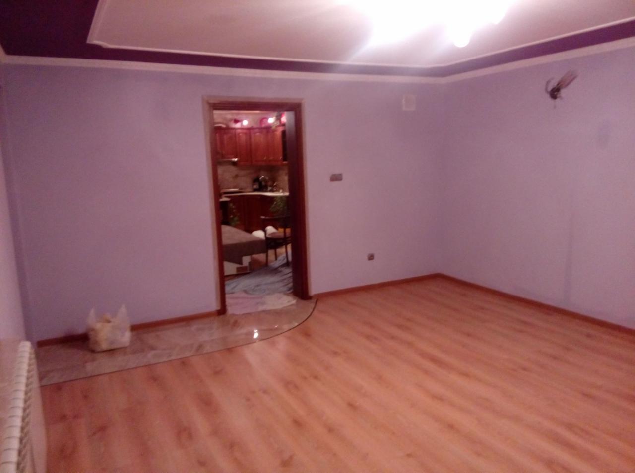 Pusty pokój po remoncie z panelami podłogowymi i świeżo pomalowanymi ścianami w odcieniach fioletu, widok na kuchnię przez otwarte drzwi.