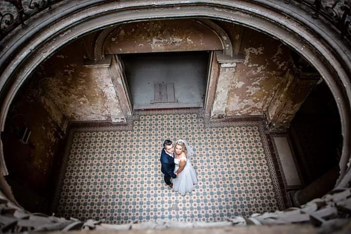 Para młoda w objęciach, sfotografowana z góry w okrągłym atrium z dekoracyjną, wzorzystą podłogą i łuszczącymi się ścianami.