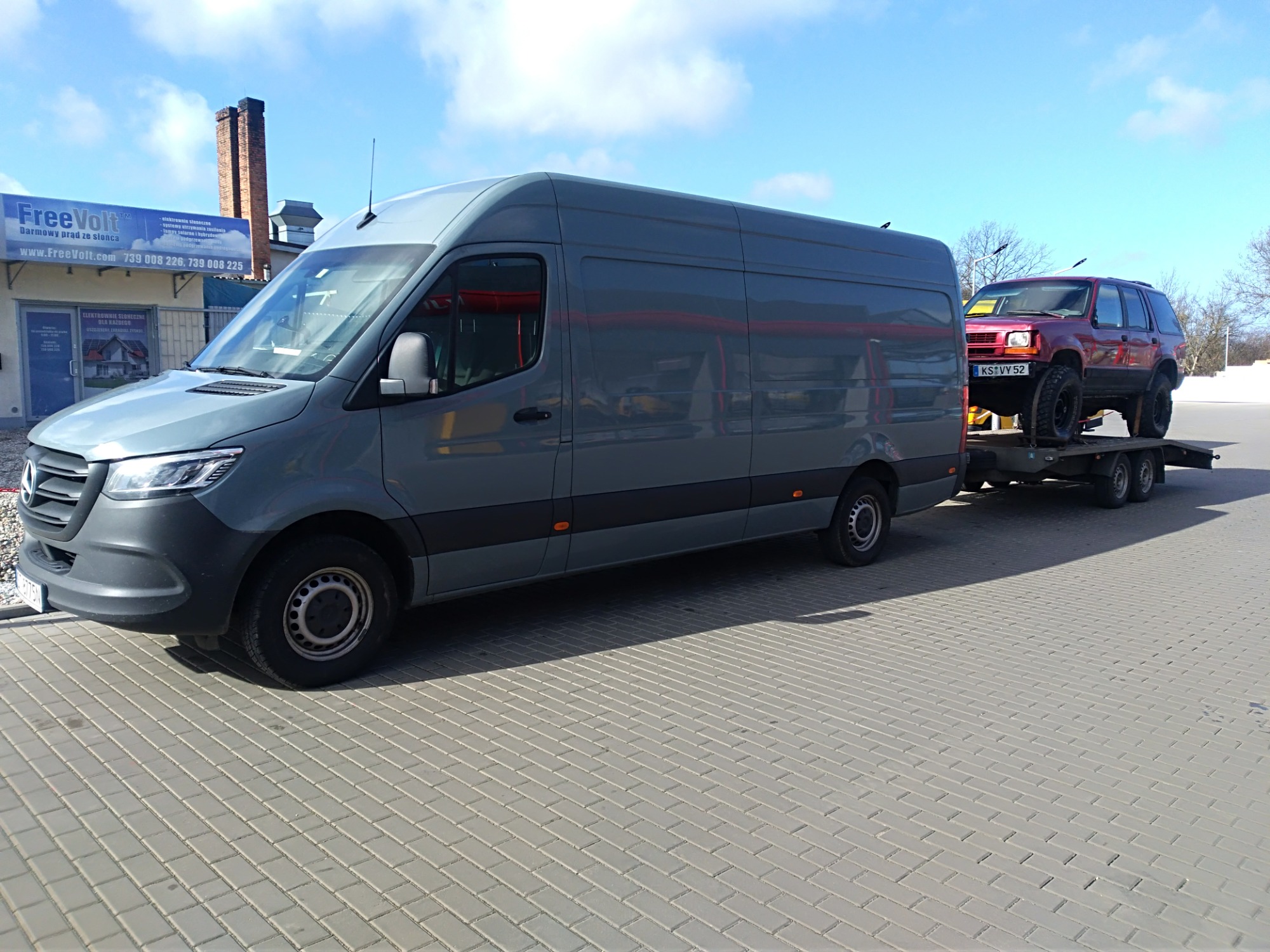 Szary bus ciągnie przyczepę z czerwonym samochodem terenowym na tle błękitnego nieba z chmurami.