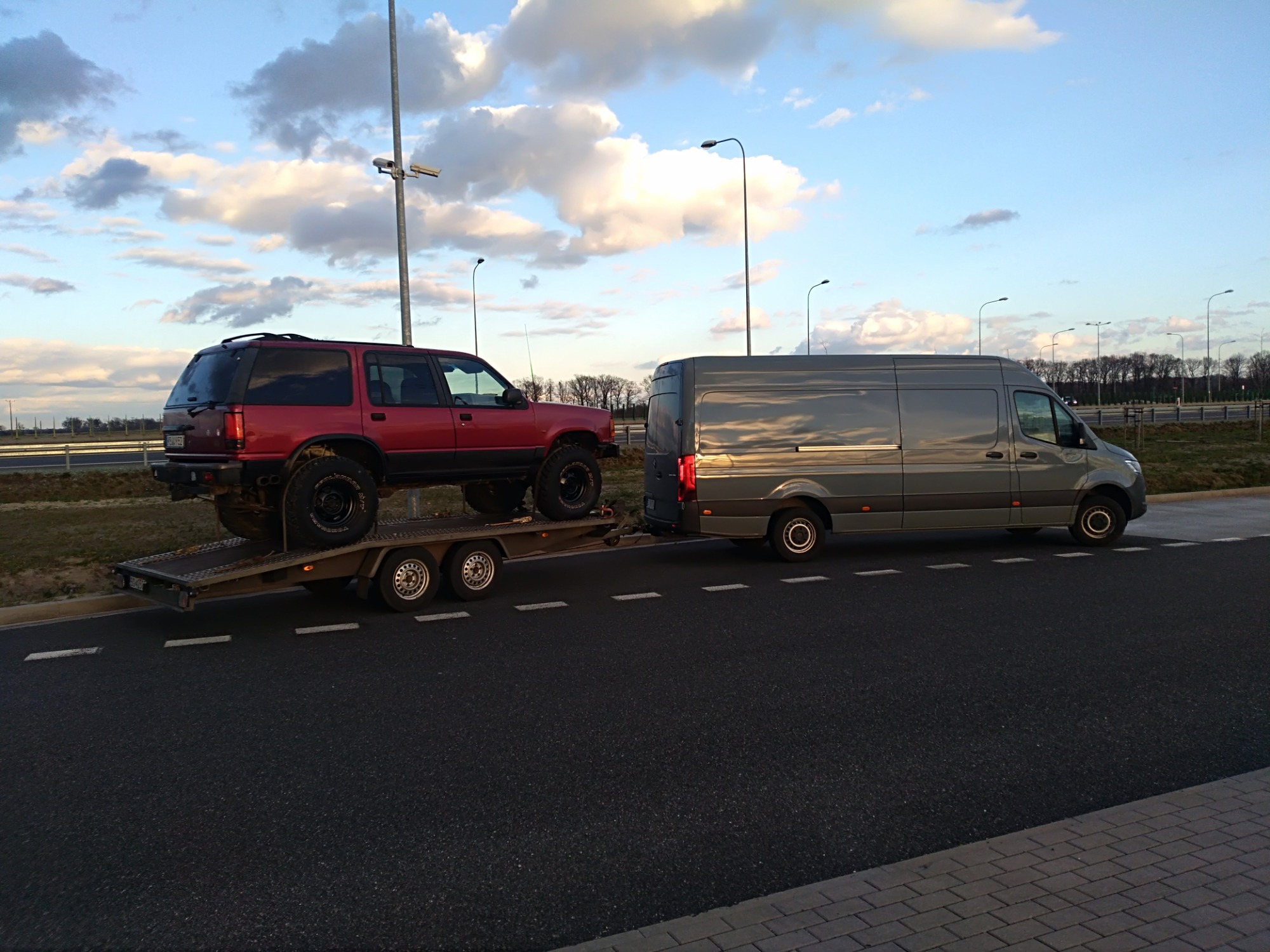 Czerwony samochód terenowy na lawecie, ciągnięty przez szary dostawczy bus na tle błękitnego nieba z chmurami i oświetlenia ulicznego.