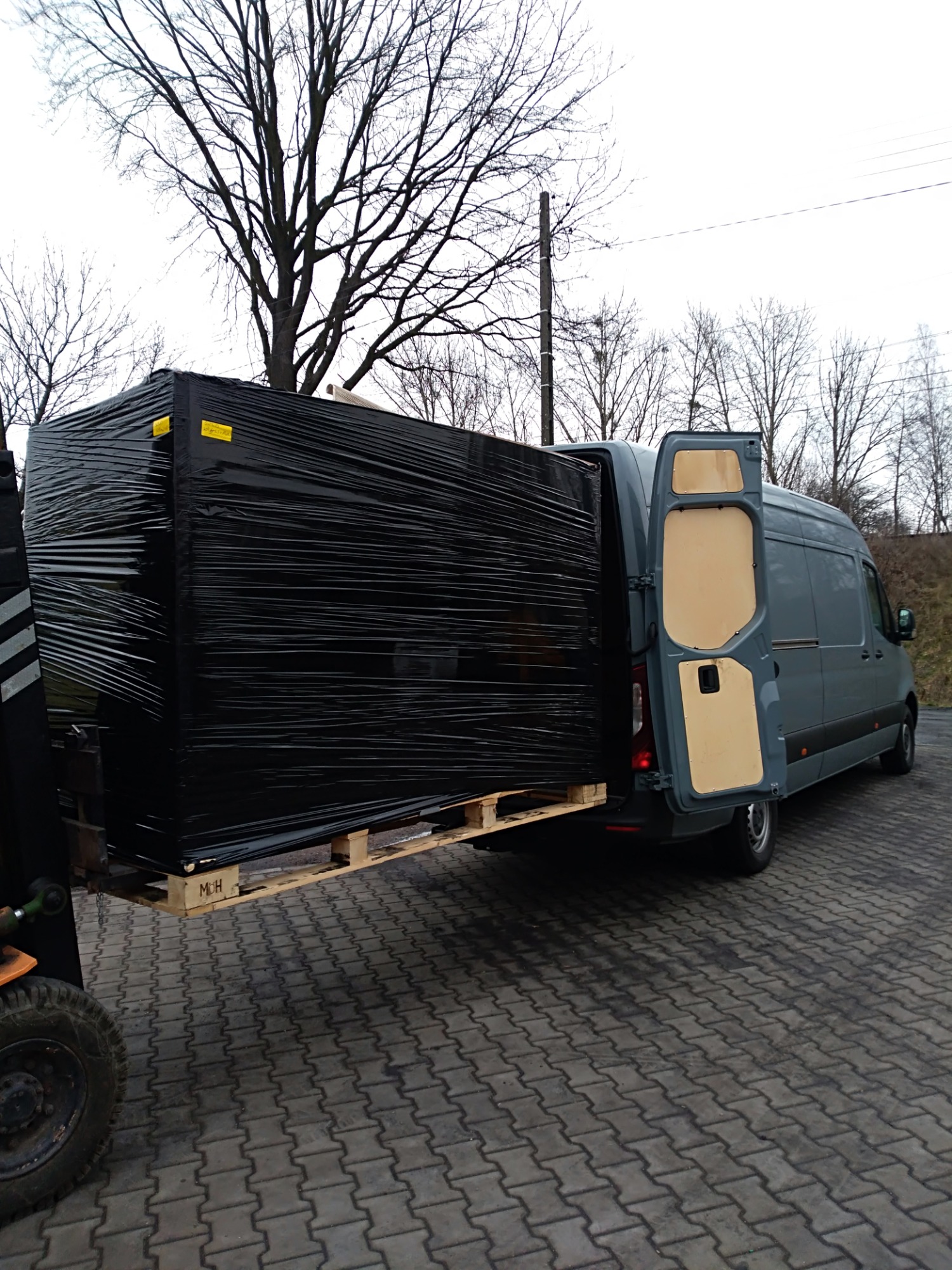 Wózek widłowy ładuje owiniętą czarną folią paletę z towarem do otwartego busa na brukowanym placu, widoczne drzewa bez liści w tle.