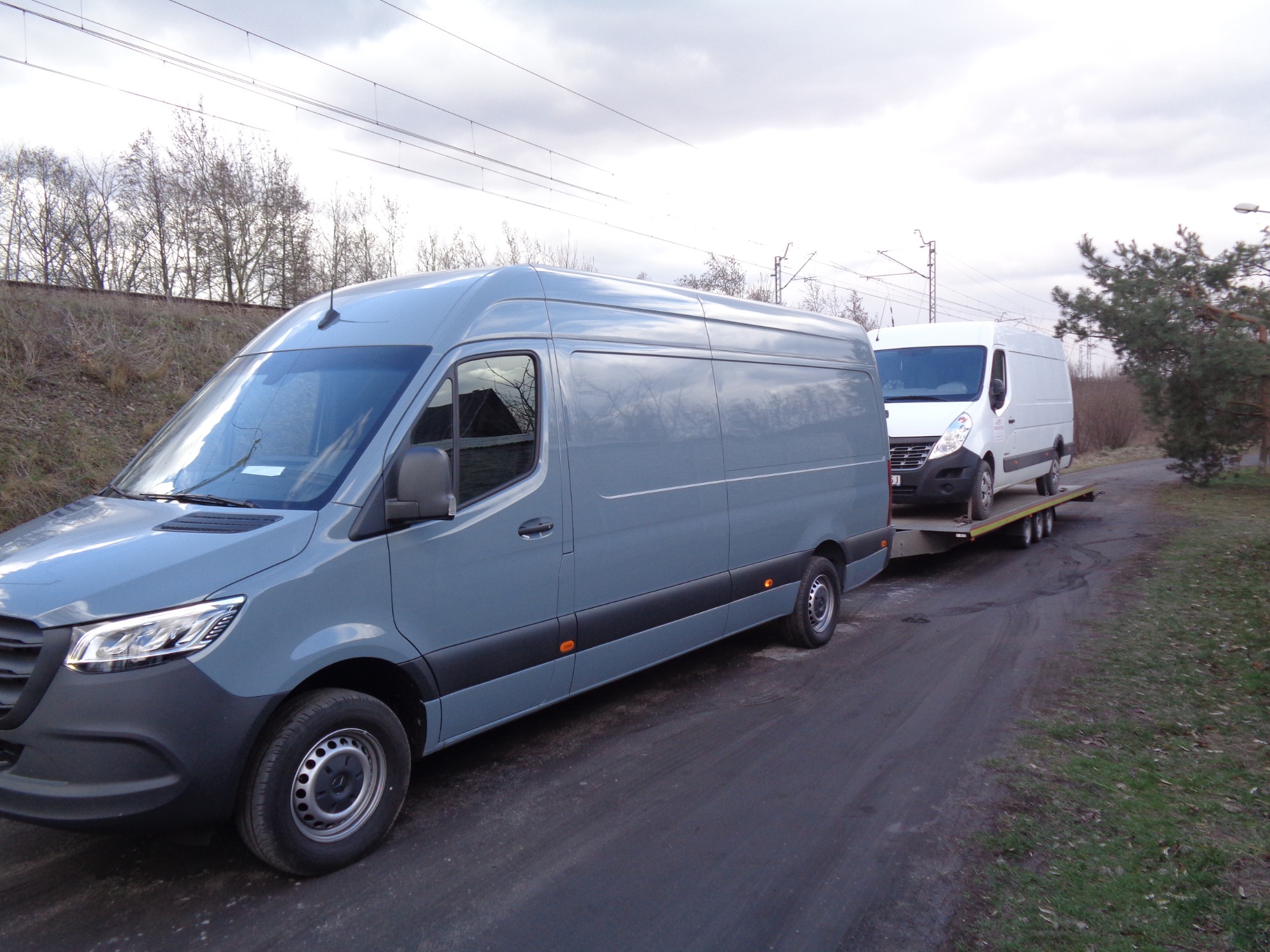 Szary bus ciągnie lawetę z białym busem na tle pochmurnego nieba i drzew bez liści.