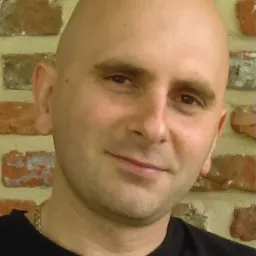 Portret mężczyzny na tle ceglanej ściany. Uśmiechnięty, łysy mężczyzna w czarnej koszulce, z delikatnym łańcuszkiem na szyi, na tle ściany z czerwonej cegły.