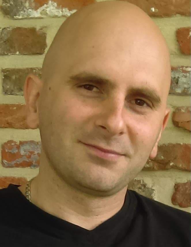 Portret mężczyzny na tle ceglanej ściany. Uśmiechnięty, łysy mężczyzna w czarnej koszulce, z delikatnym łańcuszkiem na szyi, na tle ściany z czerwonej cegły.