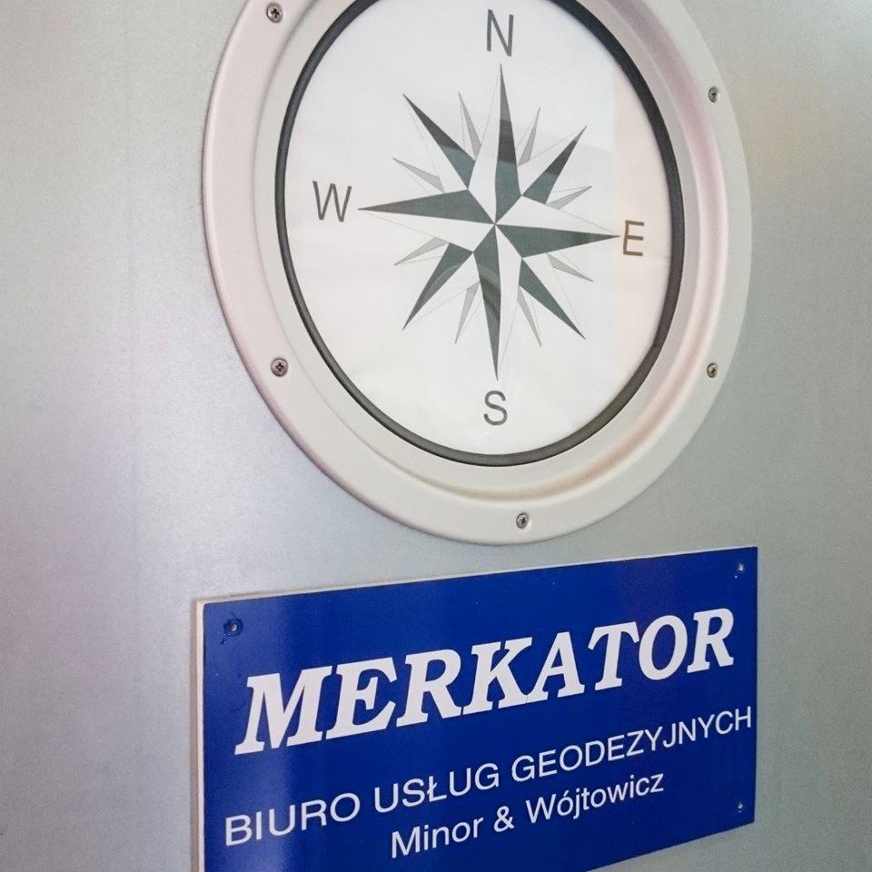 Szyld firmy 'MERKATOR Biuro Usług Geodezyjnych Minor & Wójtowicz' z dekoracyjną rozetą kompasu nad nim, przymocowany do ściany.