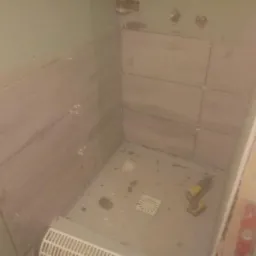 Montaż płytek imitujących drewno w kabinie prysznicowej, widoczne krzyżyki dystansowe, odpływ liniowy i wiertarka akumulatorowa.