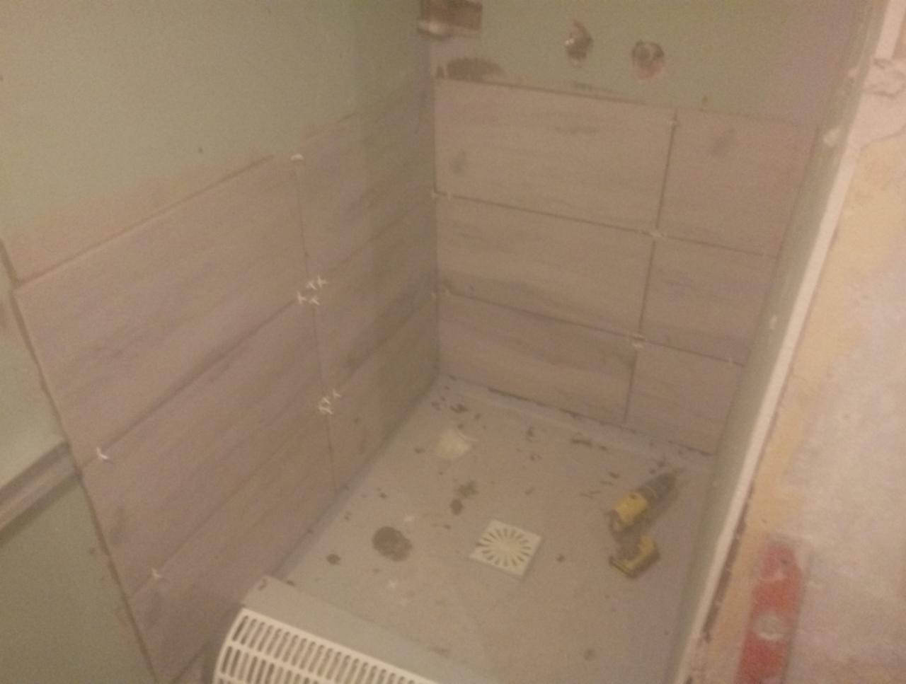 Montaż płytek imitujących drewno w kabinie prysznicowej, widoczne krzyżyki dystansowe, odpływ liniowy i wiertarka akumulatorowa.