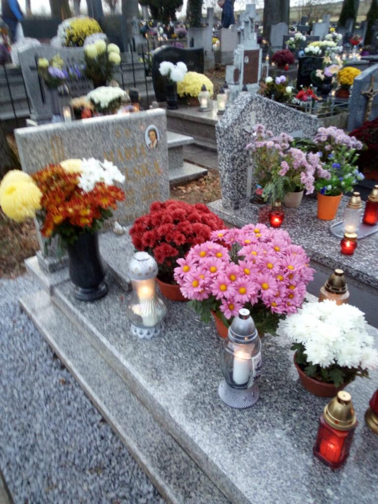 Nagrobki z kwiatami i zapalonymi zniczami w jesiennej scenerii cmentarza.