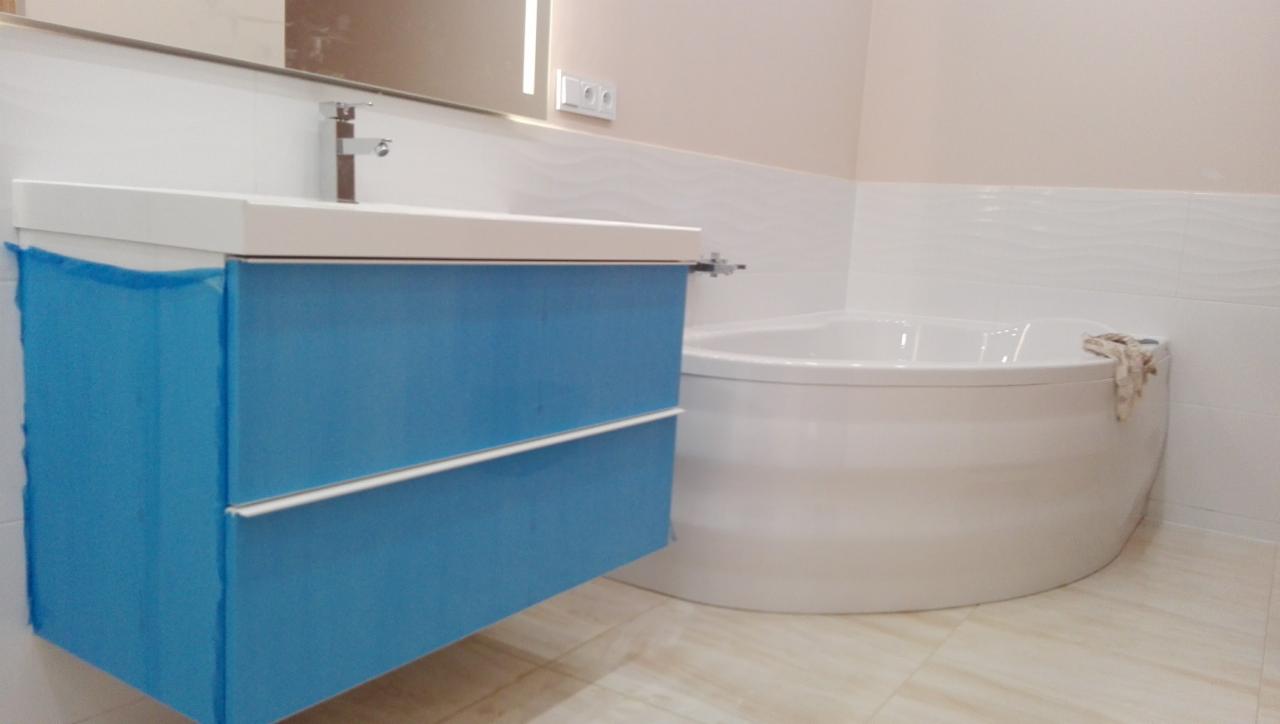 Narożna wanna i błękitna szafka pod umywalkę z folią ochronną w łazience wykończonej białymi płytkami z subtelnym wzorem fal.