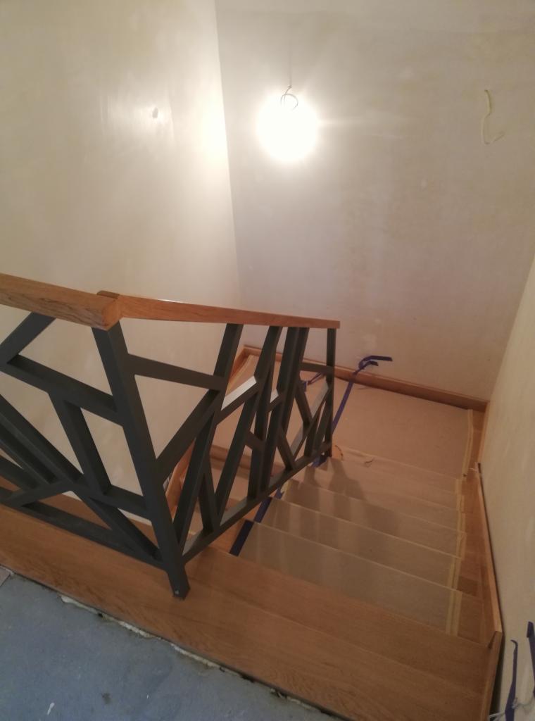 Schody z drewnianymi stopniami i balustradą o geometrycznym wzorze, widok z góry, oświetlone światłem z góry. Etap wykończeniowy, zabezpieczone taśmą malarską.