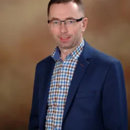 Portret mężczyzny w okularach i niebieskiej marynarce na brązowym tle. Uśmiechnięty, z krótkimi włosami, w koszuli w kratę. Profesjonalny wygląd.