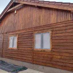 Świeżo pomalowana, brązowa, drewniana elewacja domu z zabezpieczonymi folią oknami, widoczne ślady pędzla i czarna folia ochronna na chodniku.