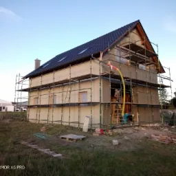 Dom jednorodzinny w trakcie prac ociepleniowych, widoczne rusztowania, pracownik z wężem natryskowym aplikujący izolację na elewację, nieotynkowana ściana szczytowa, czarne dachówki.