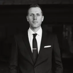 Portret mężczyzny w garniturze z krawatem i spinką, na tle ciemnej, ceglanej ściany