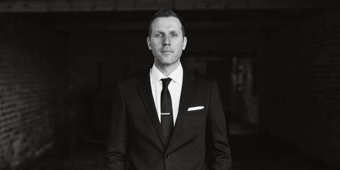 Portret mężczyzny w garniturze z krawatem i spinką, na tle ciemnej, ceglanej ściany