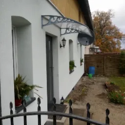 Biała elewacja domu jednorodzinnego z szarą stolarką okienną i drzwiową, ozdobnym zadaszeniem nad wejściem, lampą ścienną i anteną satelitarną, widok z poziomu ogrodzenia.